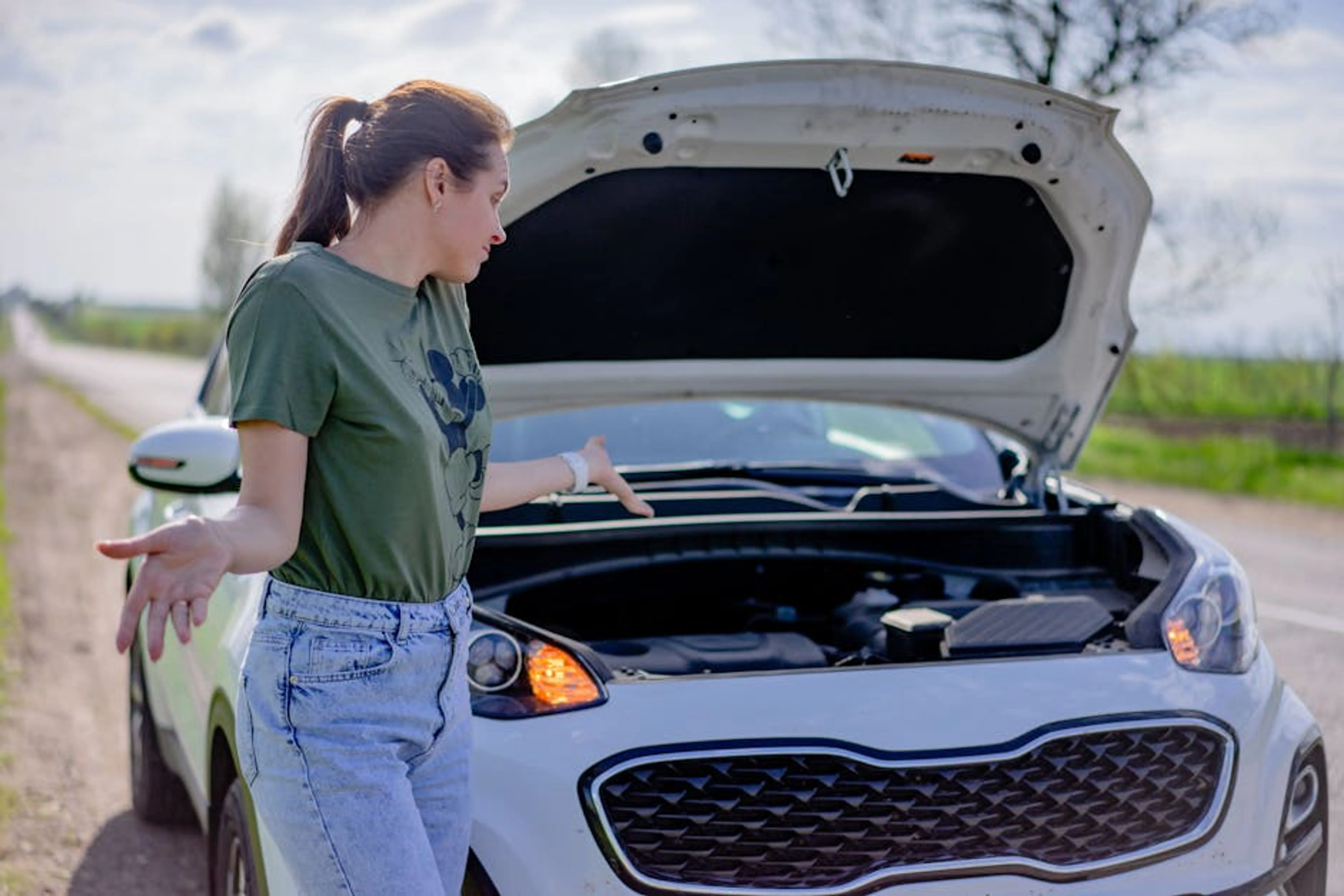 A woman looks puzzled by her broken car with an open hood on a rural road. A car pulled safely to the side of a road with hazard lights flashing, indicating a breakdown.