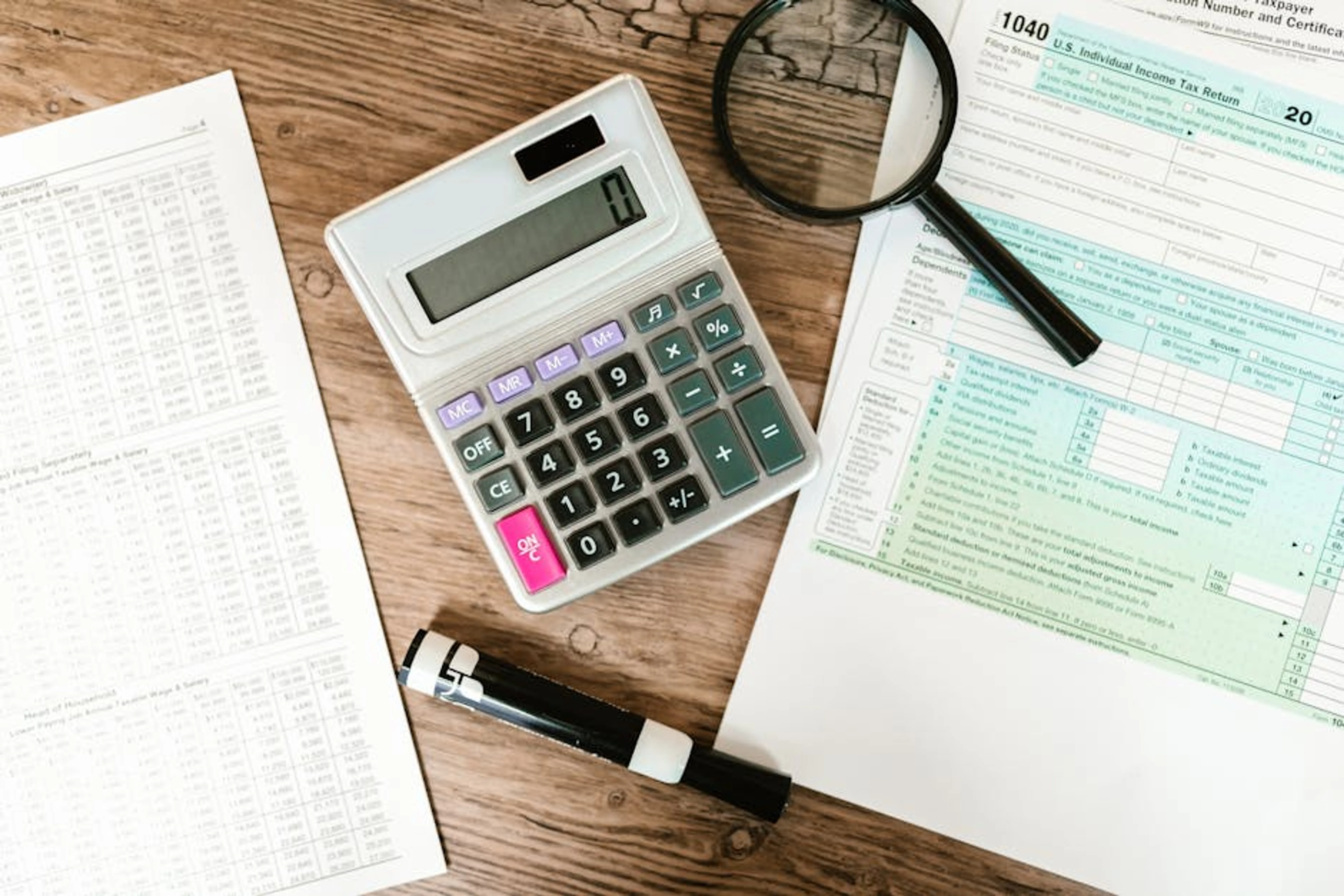 Top view of tax forms, a calculator, and a magnifying glass on a wooden surface, ideal for accounting themes. Person reviewing financial statements and debt reports on a desk