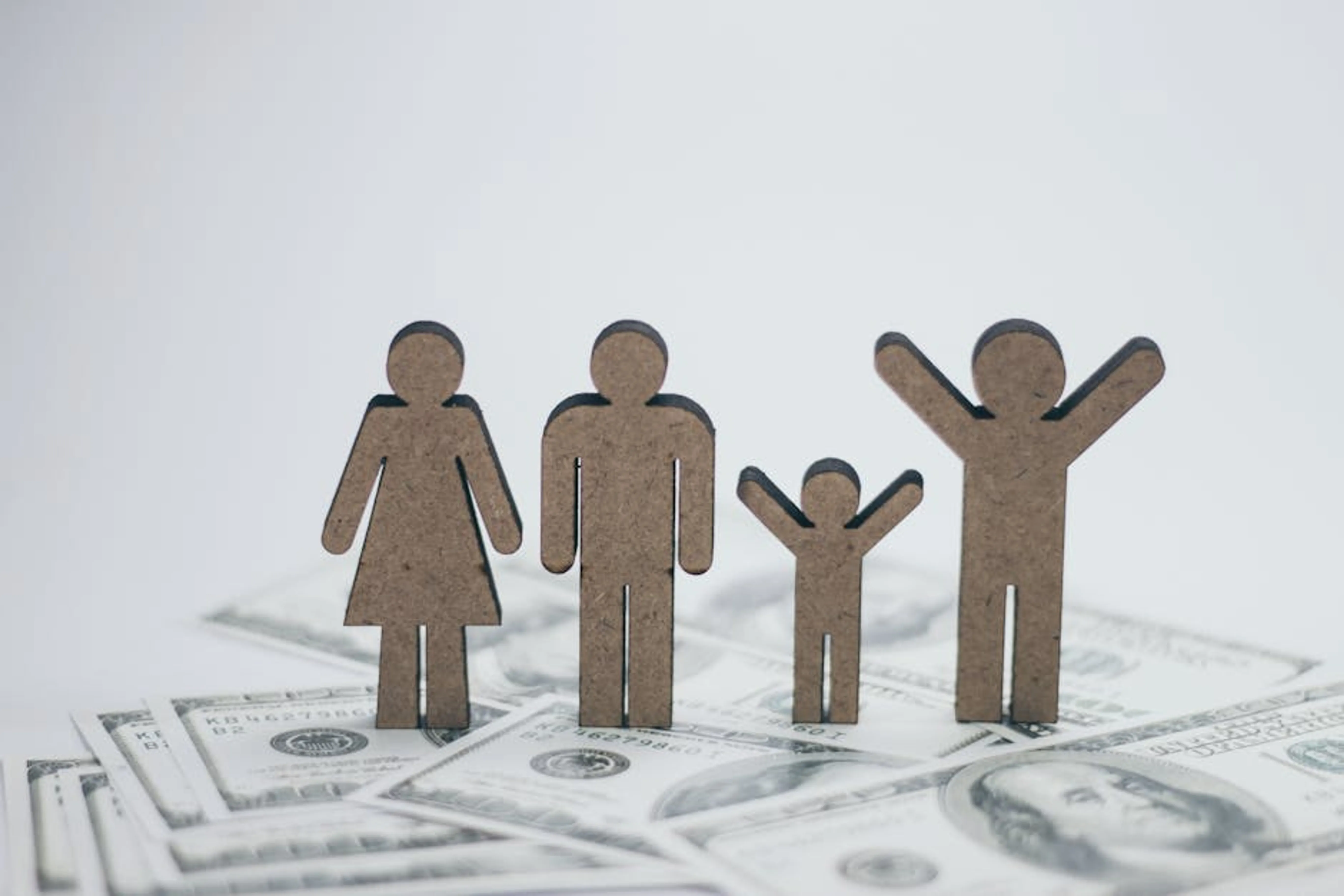 Wooden family figurines on dollar bills symbolizing financial stability. A family sitting at a kitchen table, looking concerned while reviewing bills and a laptop with financial data.