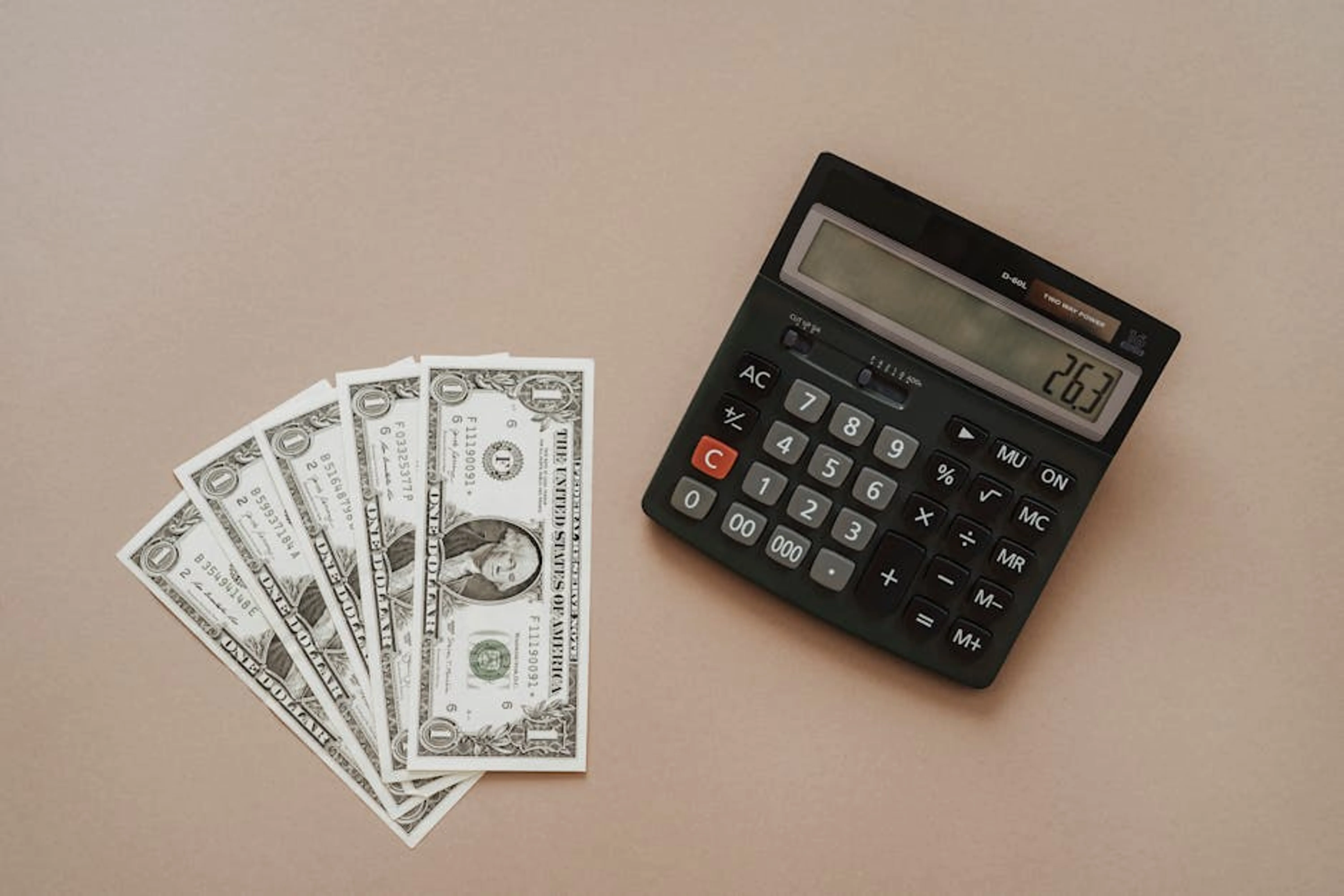A calculator displaying '26.3' beside four one-dollar bills on a beige background. Person using a financial calculator or laptop to compare refinance options and potential savings.