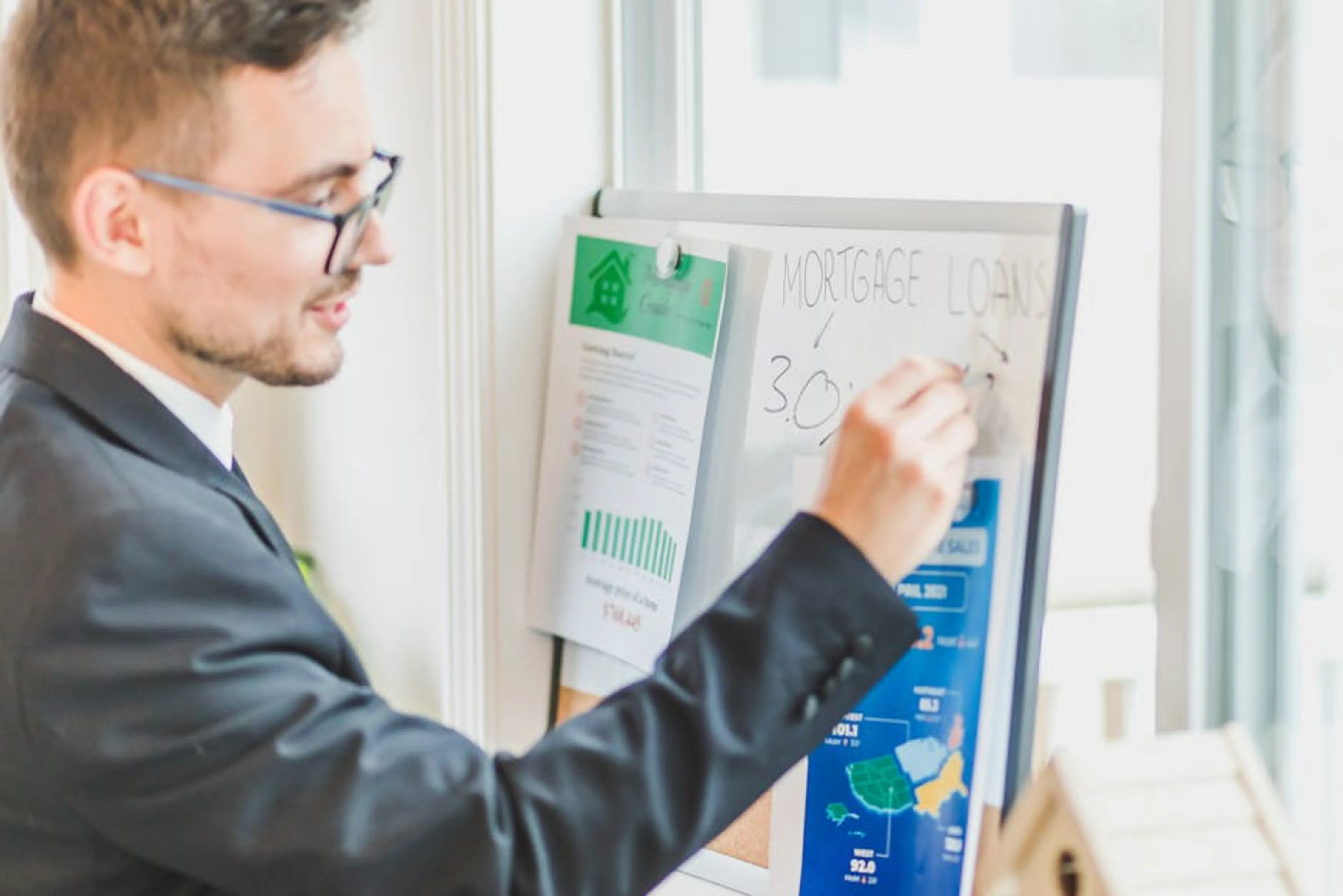 Businessman in suit writes on whiteboard displaying mortgage loan rates during a real estate meeting. Couple looking at documents and a laptop, making a thoughtful decision about their home loan or refinance.