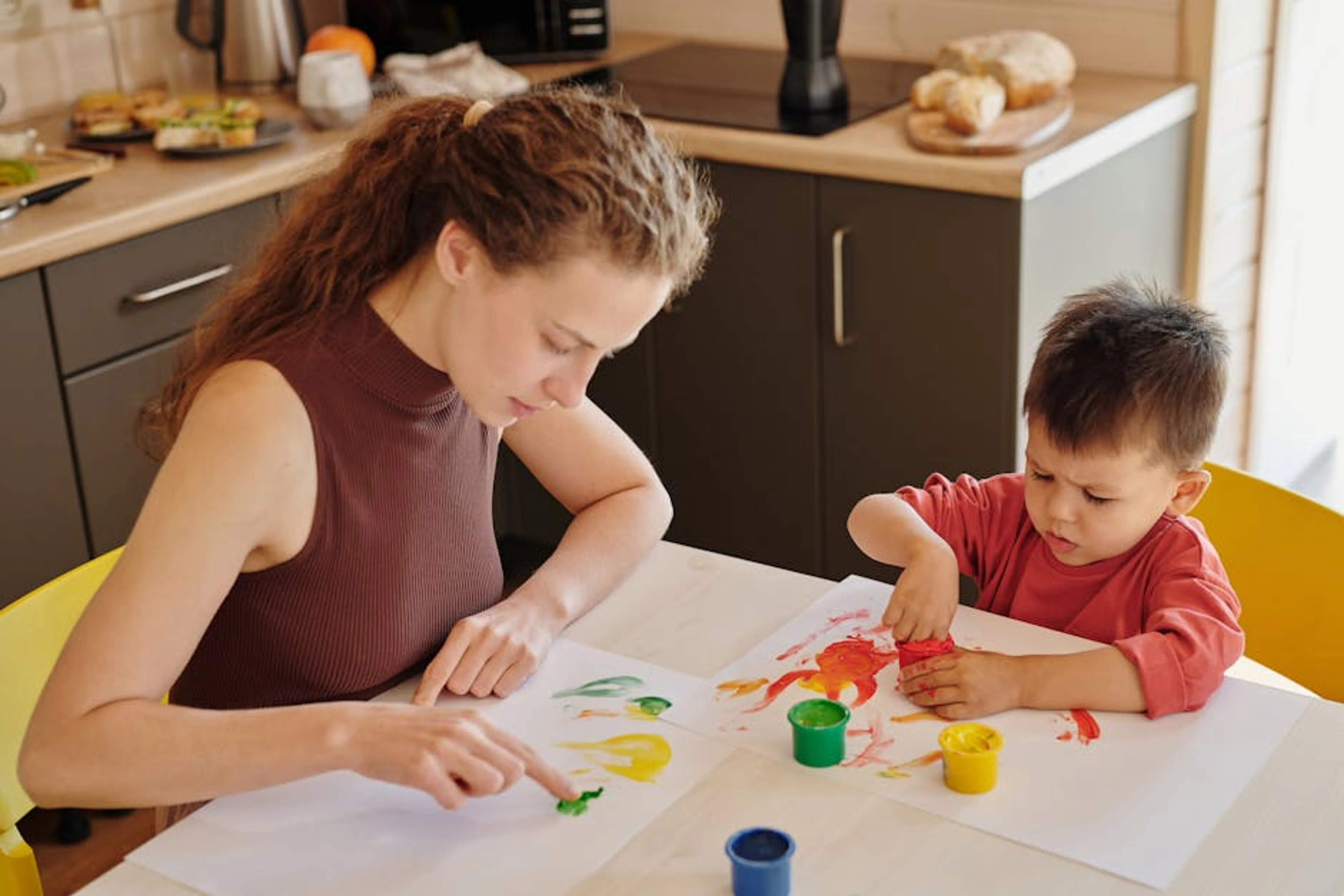 A mother and child engage in creative finger painting at a kitchen table, fostering creativity and family bonding. A toddler joyfully finger painting with bright, washable colors on a large sheet of paper, wearing a smock.