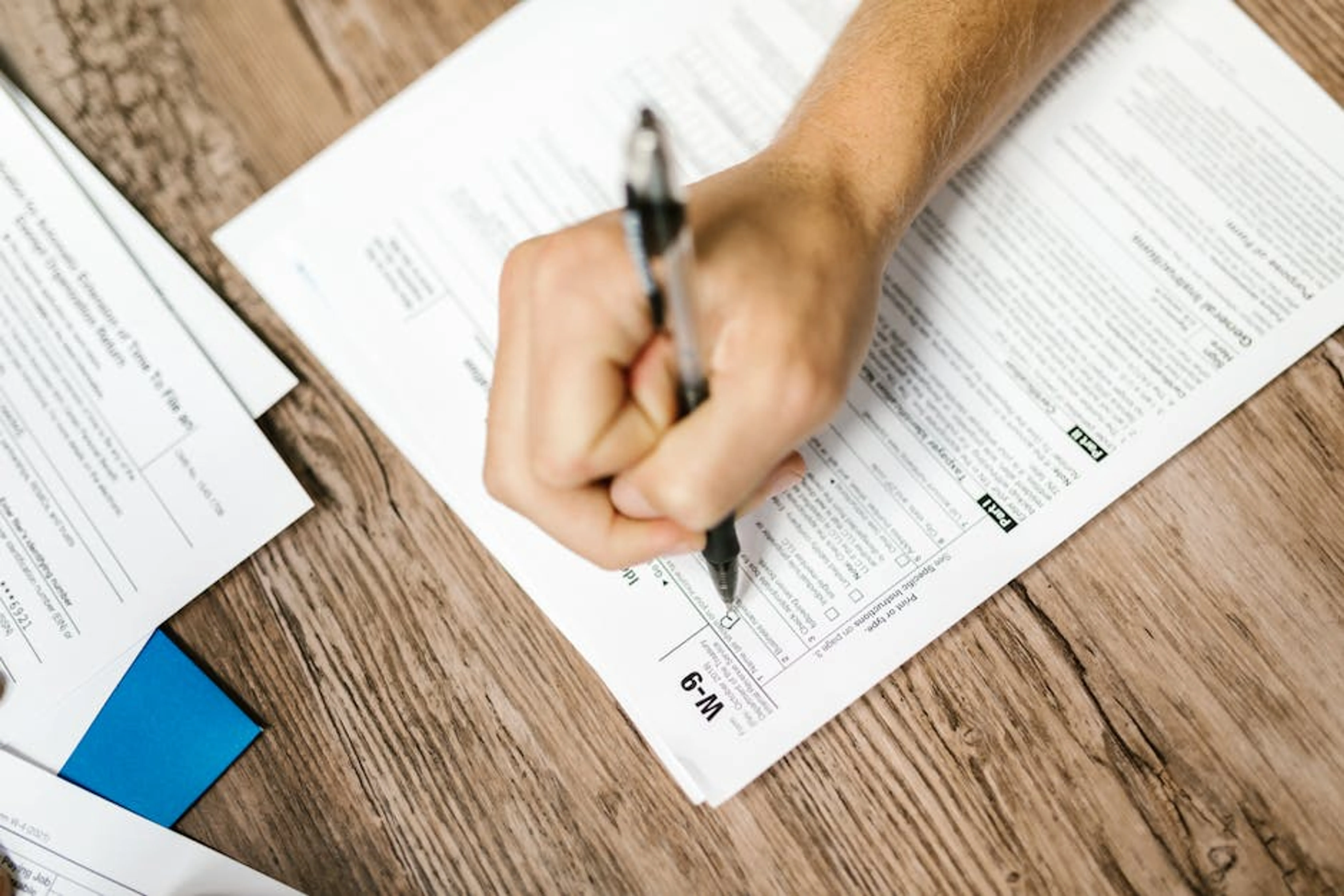 Hand writing on a W-9 tax form using a ballpoint pen on wooden desk. A person's hands filling out a workers' compensation claim form with a pen, emphasizing accuracy.